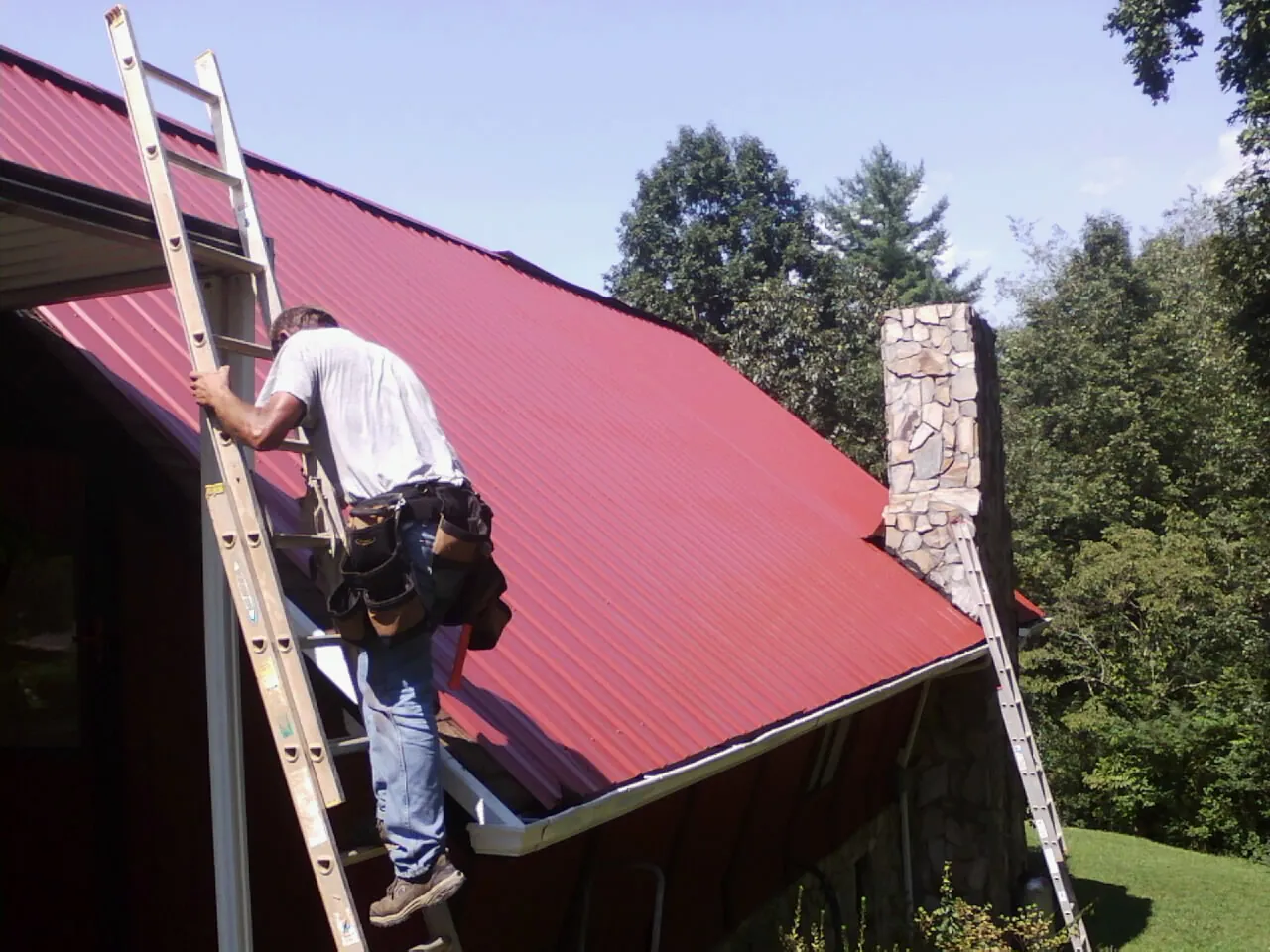 Roofer on a ladder beside a red metal roof and stone chimney — Soffit & Fascia Repair by Roofing Brothers in Piru